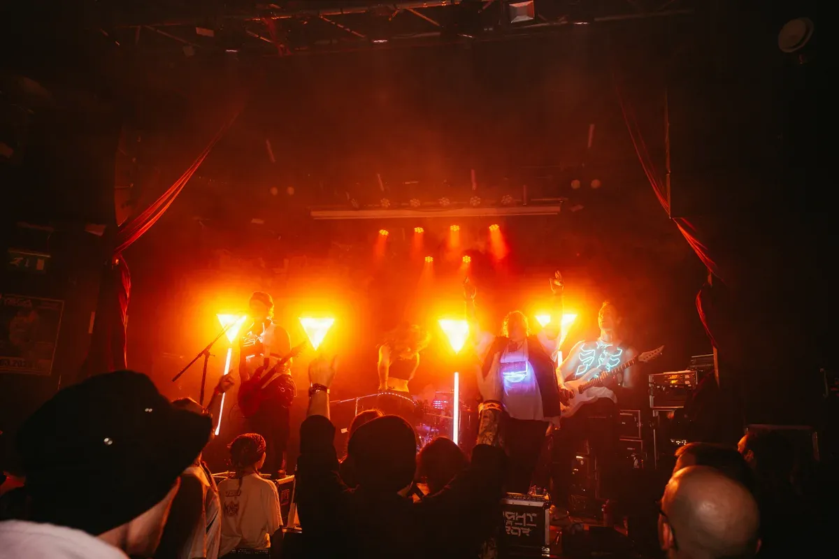 Ganze Band auf der Buehne mit orangenen Lichtern und Publikum-Silhouetten
