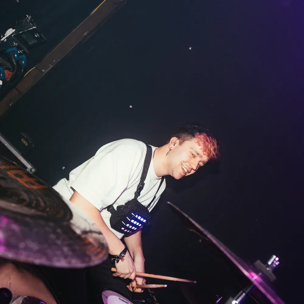 Drummer laechelt hinter seinen Becken im lila Buehnenlicht