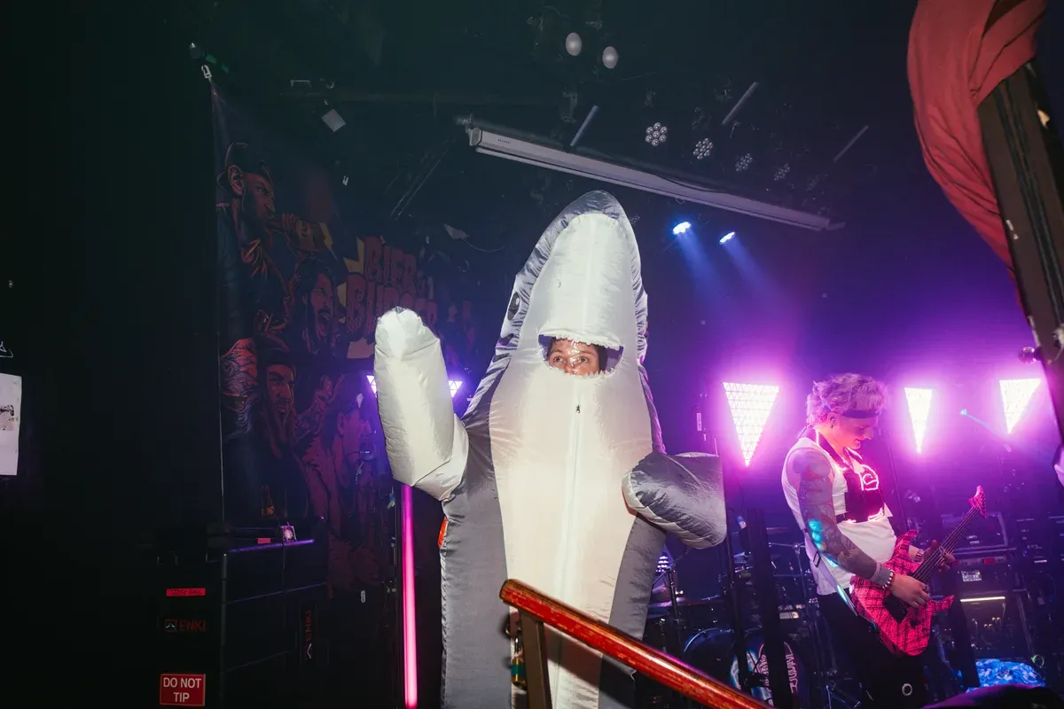 Person im Hai-Kostuem auf der Buehne neben dem Bassisten